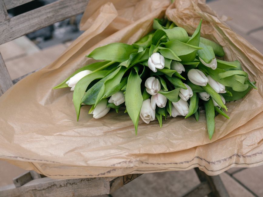 A fresh bouquet of white tulips with closed petals and vibrant green leaves, arranged in a loose cluster and wrapped in light brown tissue paper. The bouquet rests on a weathered wooden surface, with 