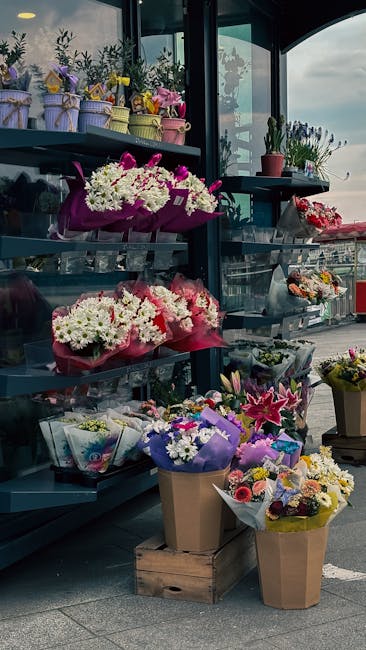 A display of floral arrangements on metal shelves outside a flower shop, featuring a variety of fresh flowers including white daisies, pink lilies, and purple statice wrapped in colorful paper and rib
