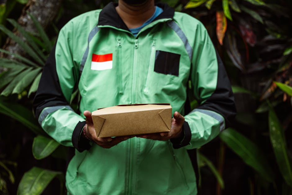 A delivery person wearing a green and black jacket with a red and white flag emblem is holding a rectangular cardboard parcel. The person is standing outdoors against a backdrop of lush green foliage,