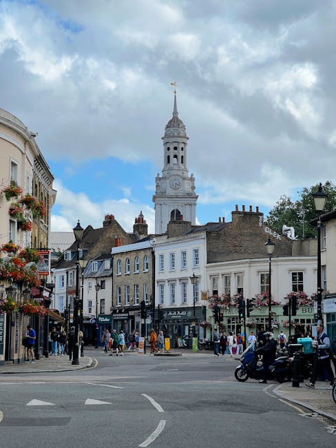 The image depicts a lively street scene in Bounds Green, with a prominent white clock tower topped by a golden weather vane against a partly cloudy sky. Surrounding the tower are classic UK-style buil