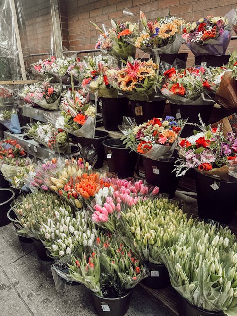 An outdoor display of numerous floral bouquets and buckets filled with fresh spring flowers, including tulips in shades of pink, white, orange, and red, arranged in black buckets and wrapped in clear 